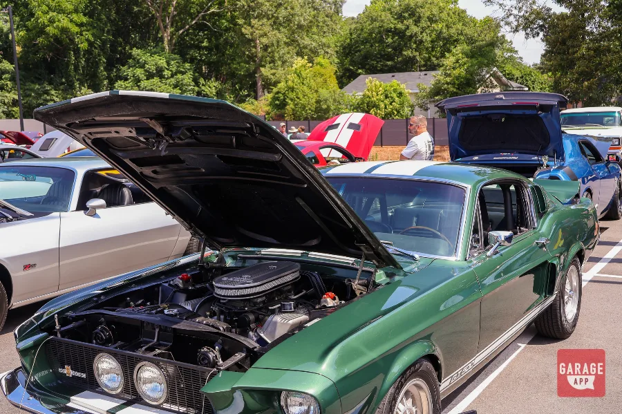 Front three-quarter view of a dark green 1968 Shelby GT500 with white racing stripes and hood open at a Cars and Coffee event.