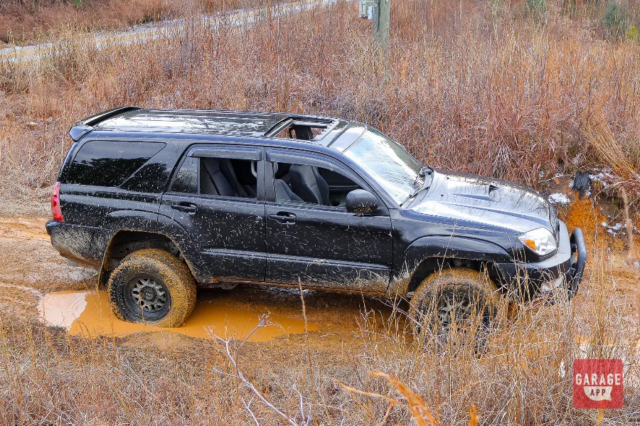 Lifted 2005 Toyota 4Runner V8 crossing a muddy water obstacle on an off-road trail surrounded by forest.