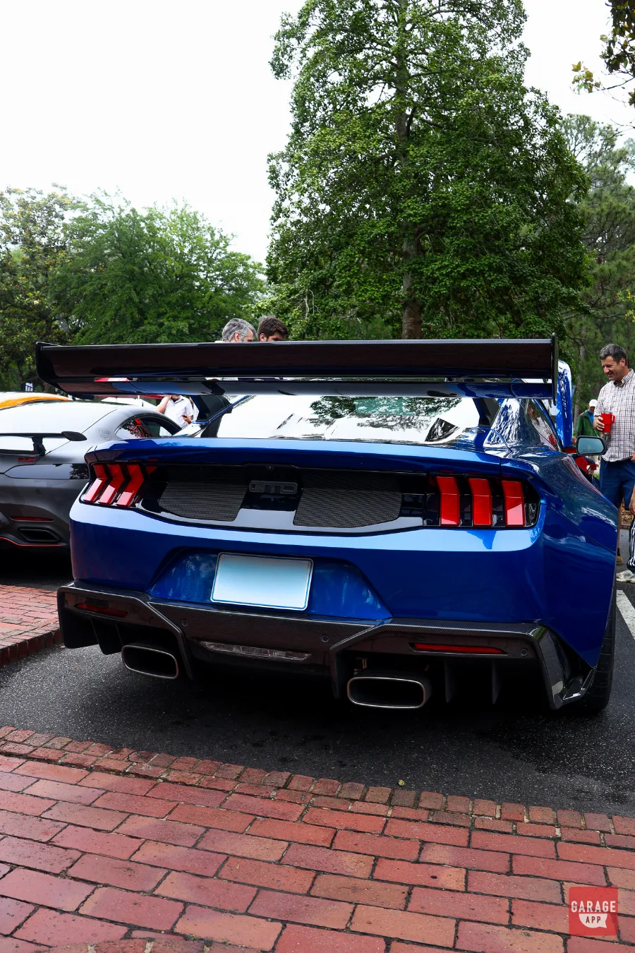 Rear view of a blue 2025 Ford Mustang GTD featuring a carbon fiber rear wing, quad exhaust outlets, aggressive diffuser, and GTD badging at a Cars and Coffee event.