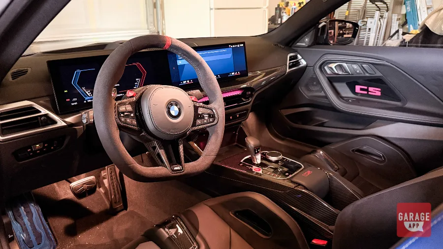 2026 BMW M2 CS driver cockpit with Alcantara steering wheel, carbon fiber trim, illuminated CS door panel, and digital displays.