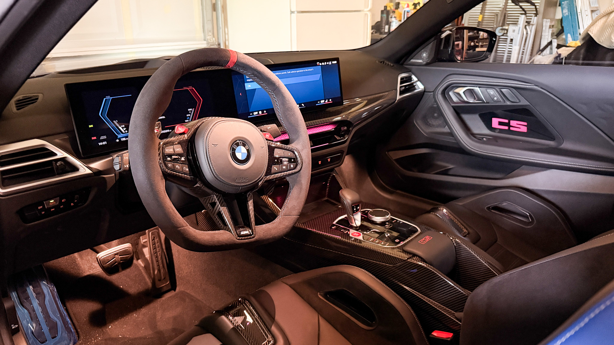 2026 BMW M2 CS driver cockpit with Alcantara steering wheel, carbon fiber trim, illuminated CS door panel, and digital displays.