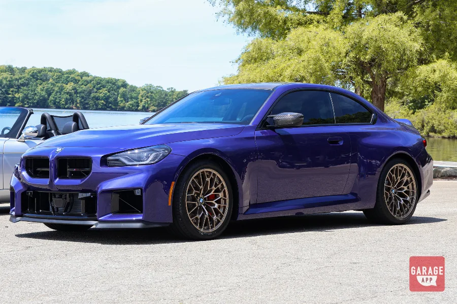 2026 BMW M2 CS in Velvet Blue parked beside a lake, showing the front three-quarter view and bronze forged wheels.