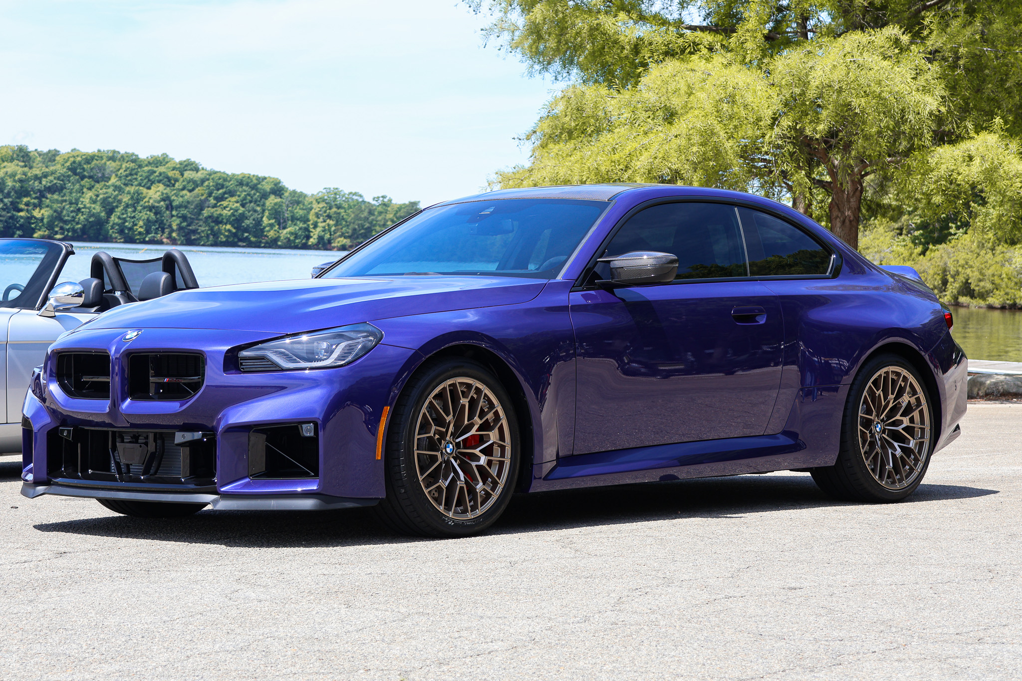 2026 BMW M2 CS in Velvet Blue parked beside a lake, showing the front three-quarter view and bronze forged wheels.