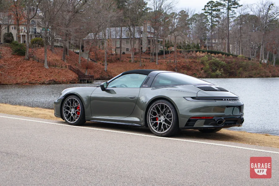 Rear three-quarter view of a 992.1 Porsche 911 Targa 4S in Aventurine Green Metallic parked beside a lakeside road, showcasing the iconic Targa roof.