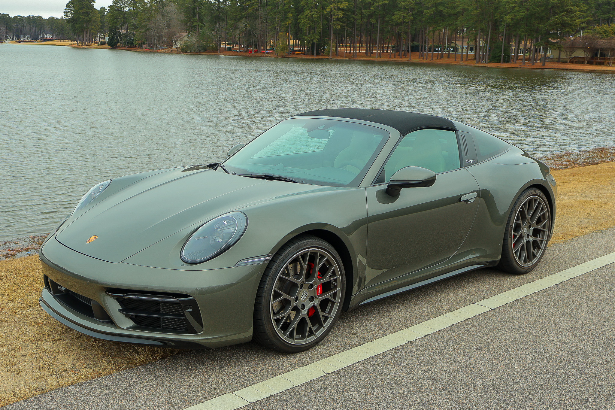 High front three-quarter view of a 992.1 Porsche 911 Targa 4S in Aventurine Green Metallic parked beside a scenic lakeside road.
