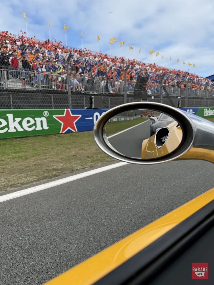 Yellow Spyker C8 side mirror reflecting car behind packed orange-clad grandstand at Zandvoort