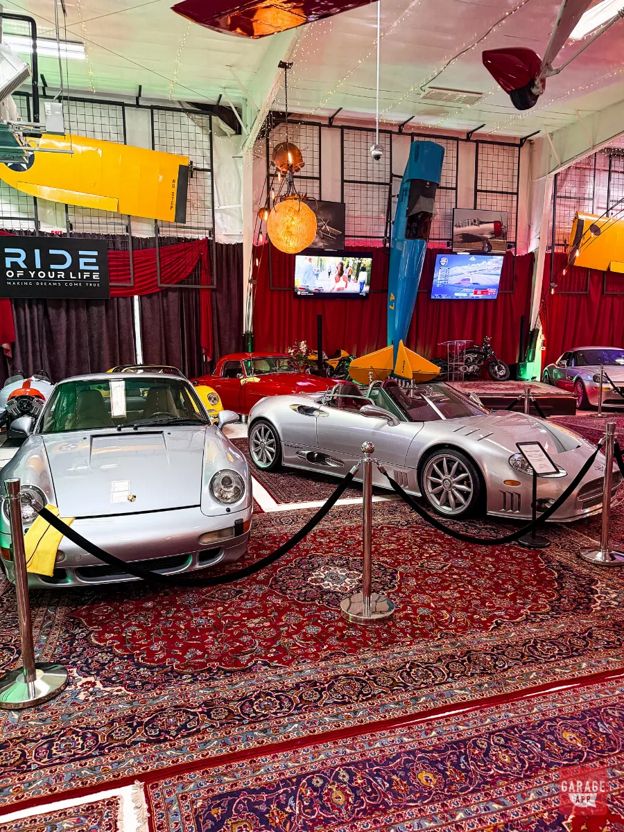 Silver Porsche 911 and silver Spyker C8 roped off inside vintage airplane-themed event venue